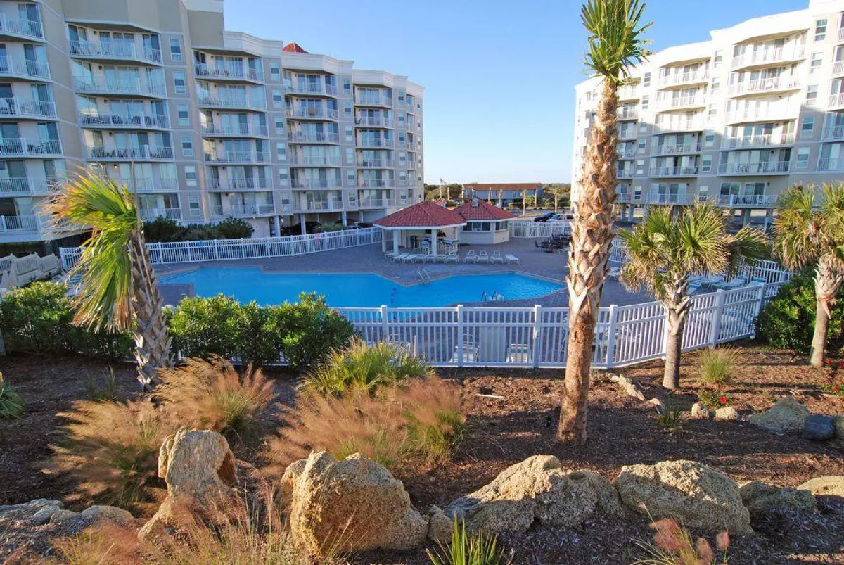 A tranquil outdoor swimming pool surrounded by palm trees and landscaped gardens, with condo buildings in the background. The pool is covered, suggesting year-round use, and lounge areas are visible for relaxation.