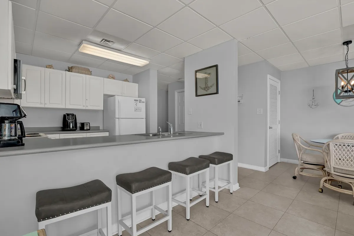 The image spotlights a kitchen island with seating, sleek white cabinetry, and a dining area with rattan chairs in the background. The inviting space is set for both casual and festive gatherings, blending kitchen and dining functionality.