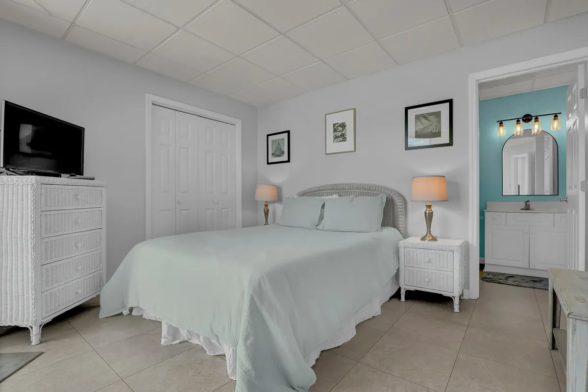 A beautifully styled bedroom with a plush bed dressed in light blue bedding, complemented by white wicker furniture, a decorative vanity area with a mirror, and tasteful wall art. Soft evening light creates a serene atmosphere.