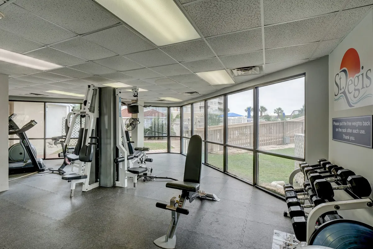 An inviting on-site fitness center with exercise machines, a large rack of weights, and generous natural light streaming through expansive windows. The room encourages active self-care, offering space for personal training or group fitness sessions.