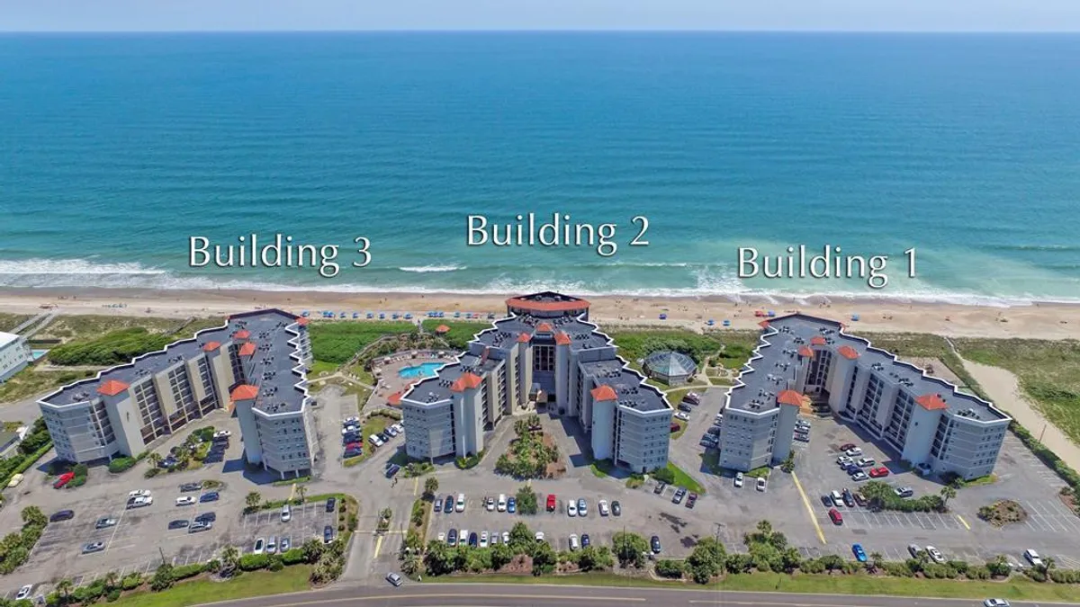 A wide-angle exterior shot capturing the resort property, including three buildings, a sparkling swimming pool, and the beach just beyond. The image showcases the proximity of resort amenities to the shoreline, with space for families to gather and relax.