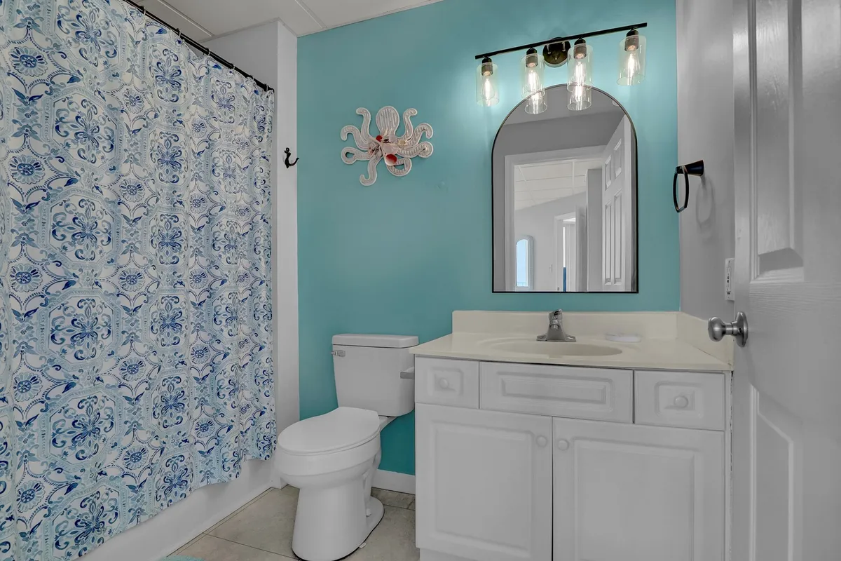 A bright and inviting bathroom with a blue shower curtain featuring intricate patterns, a round mirror above the sink, and whimsical octopus wall decor. The space feels fresh and energizing, ideal for morning routines or post-beach relaxation.