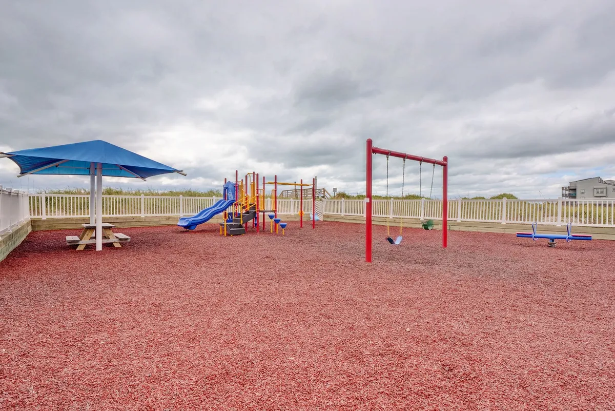A cheerful playground area featuring modern play equipment, swings, a picnic table, and a shaded structure on a sunny day. The space is designed for children to play and families to gather for snacks or picnics between outings.