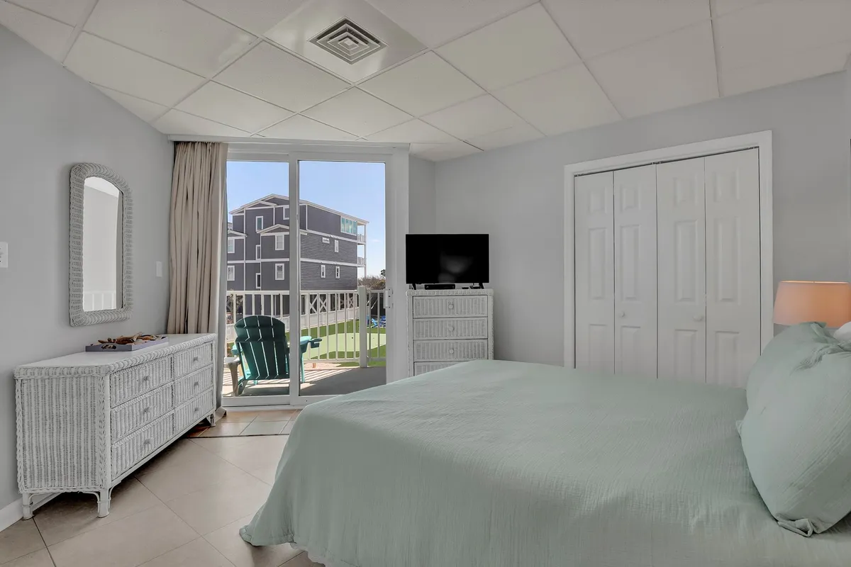 A serene and comfortable bedroom featuring a bed, sliding glass door, dresser, and a television. Natural light fills the room, suggesting a welcoming retreat with convenient amenities.