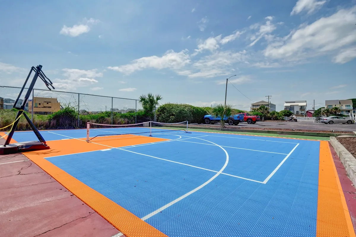 An outdoor sports court area featuring a colorful basketball court, a tennis net, and plenty of open space for group activities. The bright, energetic scene suggests friendly competitions and group recreation under the sun.