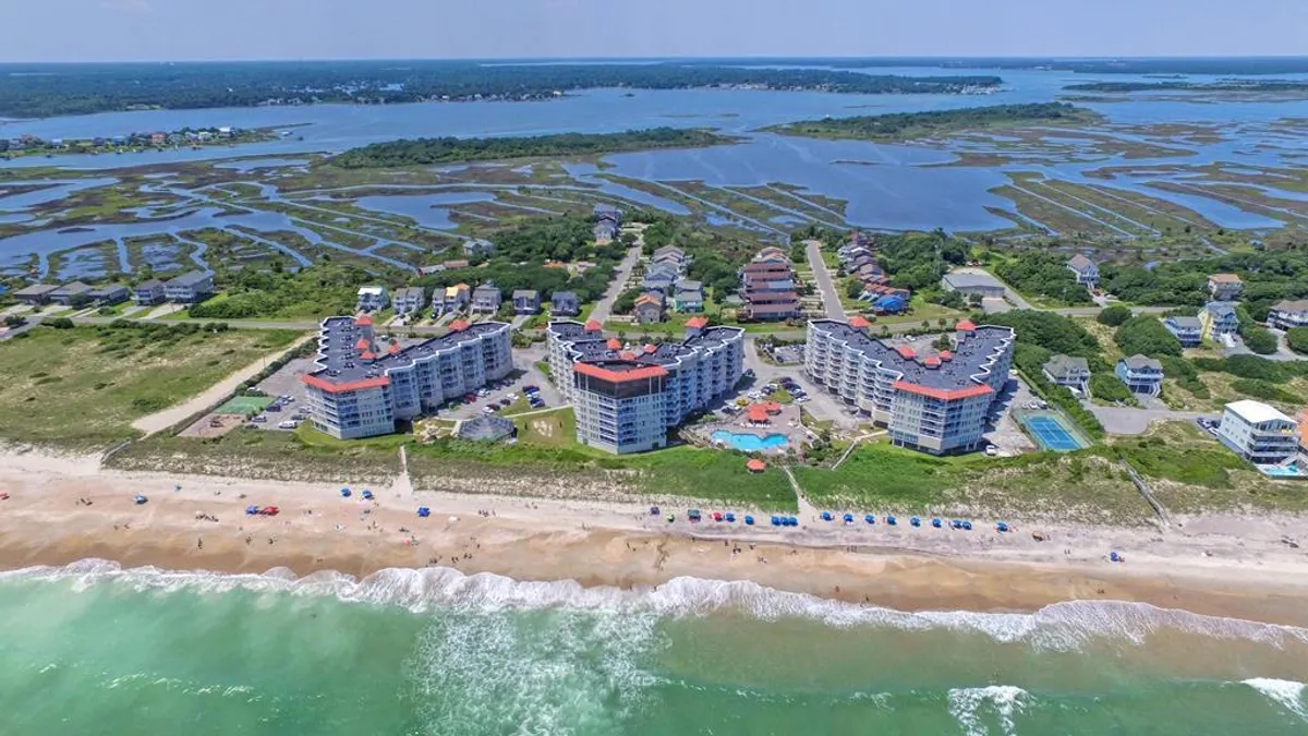 A wide-angle exterior shot featuring the resort’s beachfront with umbrellas, marshlands, the main resort buildings, and a sparkling swimming pool. The scene is vibrant, with people sunbathing, swimming, and enjoying a variety of recreational activities along the shore.