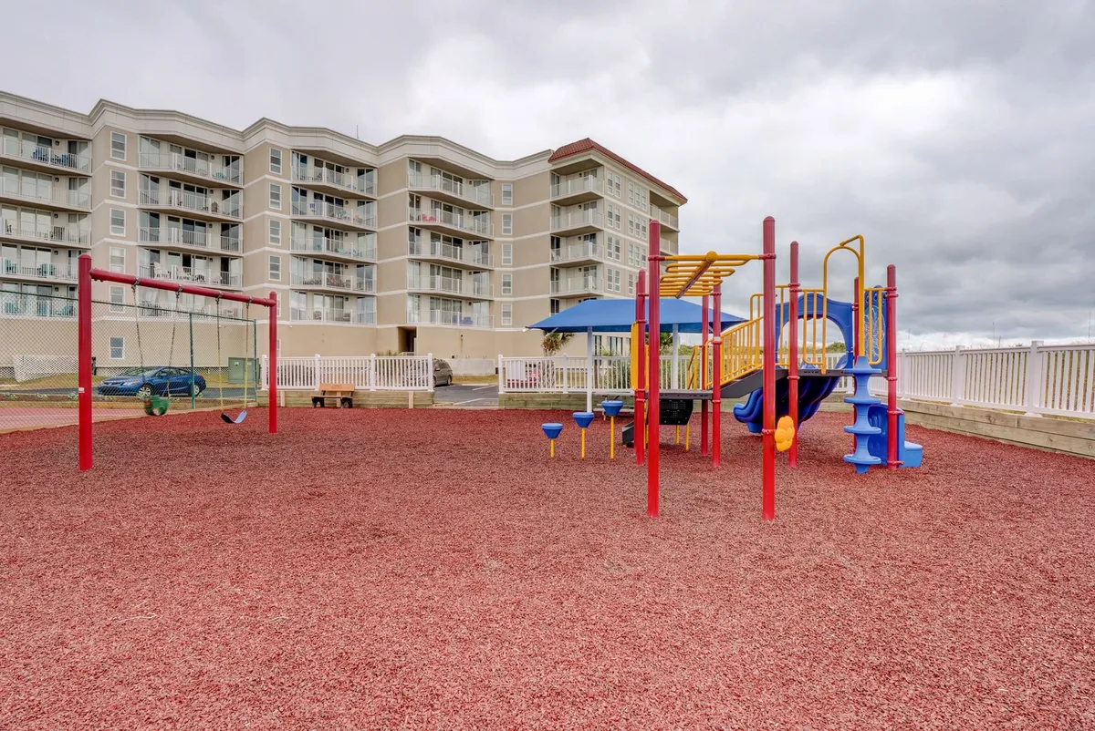 A vibrant playground scene with colorful equipment, swings, and a red rubber ground cover. Families and children can be imagined enjoying the space, and the building in the background situates the playground within the residential context.