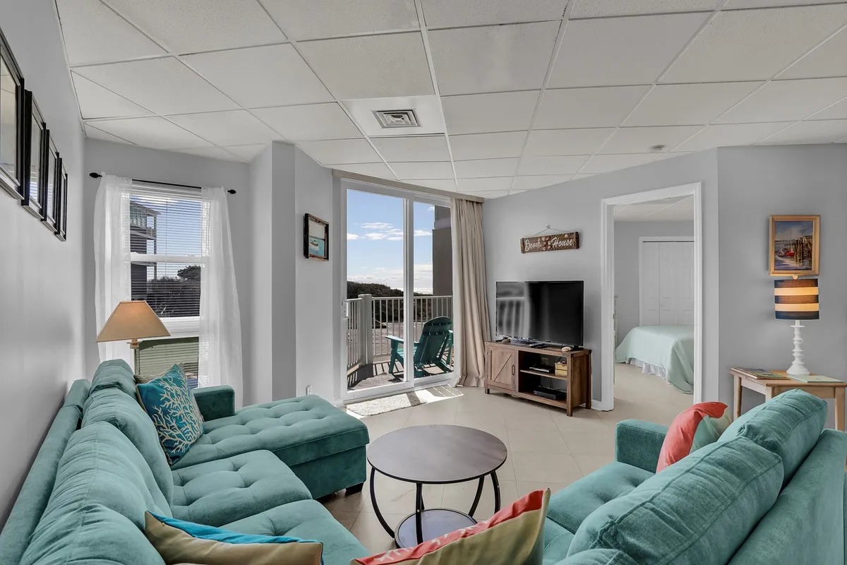The image shows a bright, open living area with a turquoise sectional sofa, a round coffee table, and large windows that pour in natural light. A television unit sits on the wall, and the view extends toward a breezy balcony, reinforcing the theme of coastal comfort and togetherness.