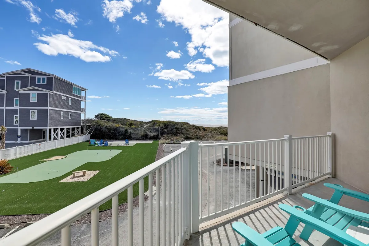 The image shows a condo balcony overlooking a lush green lawn with a clear blue sky above. The scene includes glimpses of nearby houses, suggesting a peaceful neighborhood setting. The balcony appears inviting, perfect for relaxing, enjoying a morning coffee, or spending time with friends and family while taking in the fresh coastal air.